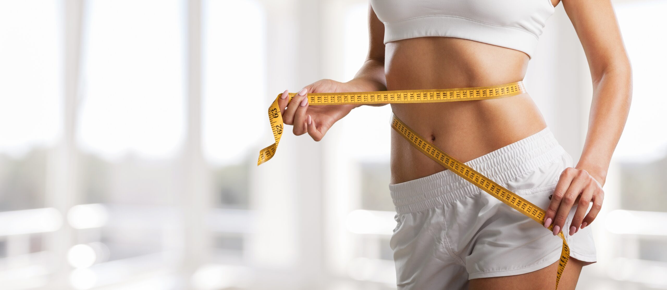 Woman in white athletic wear measuring her waist with a yellow tape measure in a bright, sunlit room