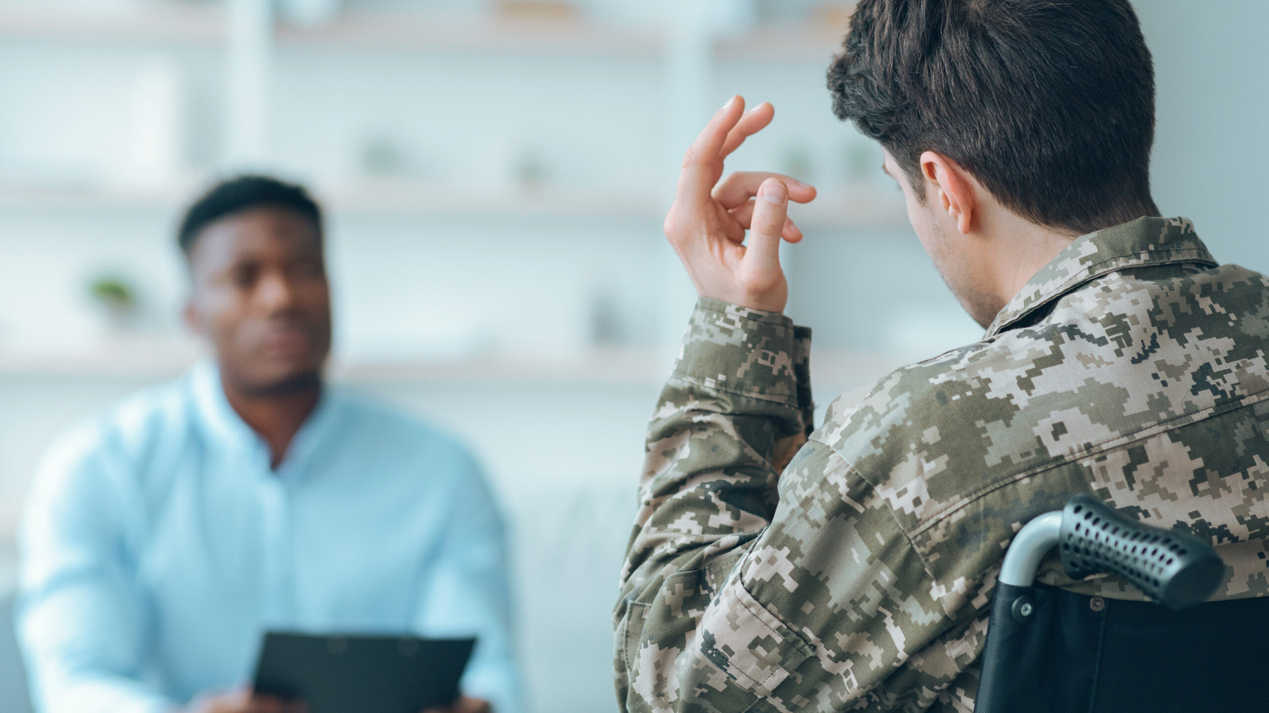 Over-the-shoulder view of a veteran in military camouflage uniform gesturing during a medical disability examination with a clinician holding a clipboard.
